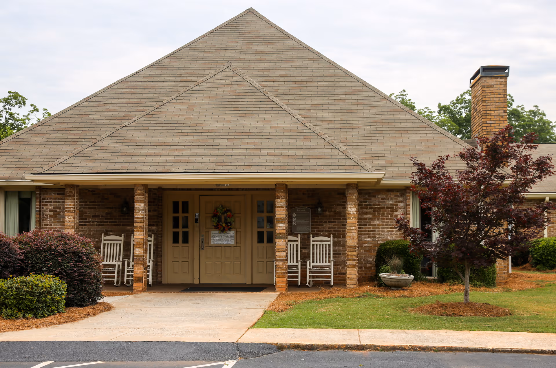 Brick single-story building front with a covered porch, double doors decorated with a wreath, rocking chairs, and landscaped shrubs and lawn.