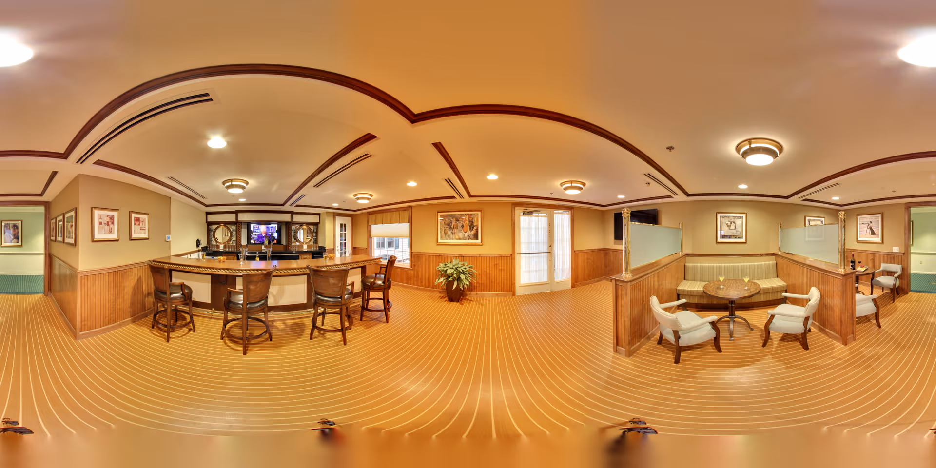 Panoramic view of a senior living facility communal lounge with a bar, bar stools, round tables and upholstered chairs.