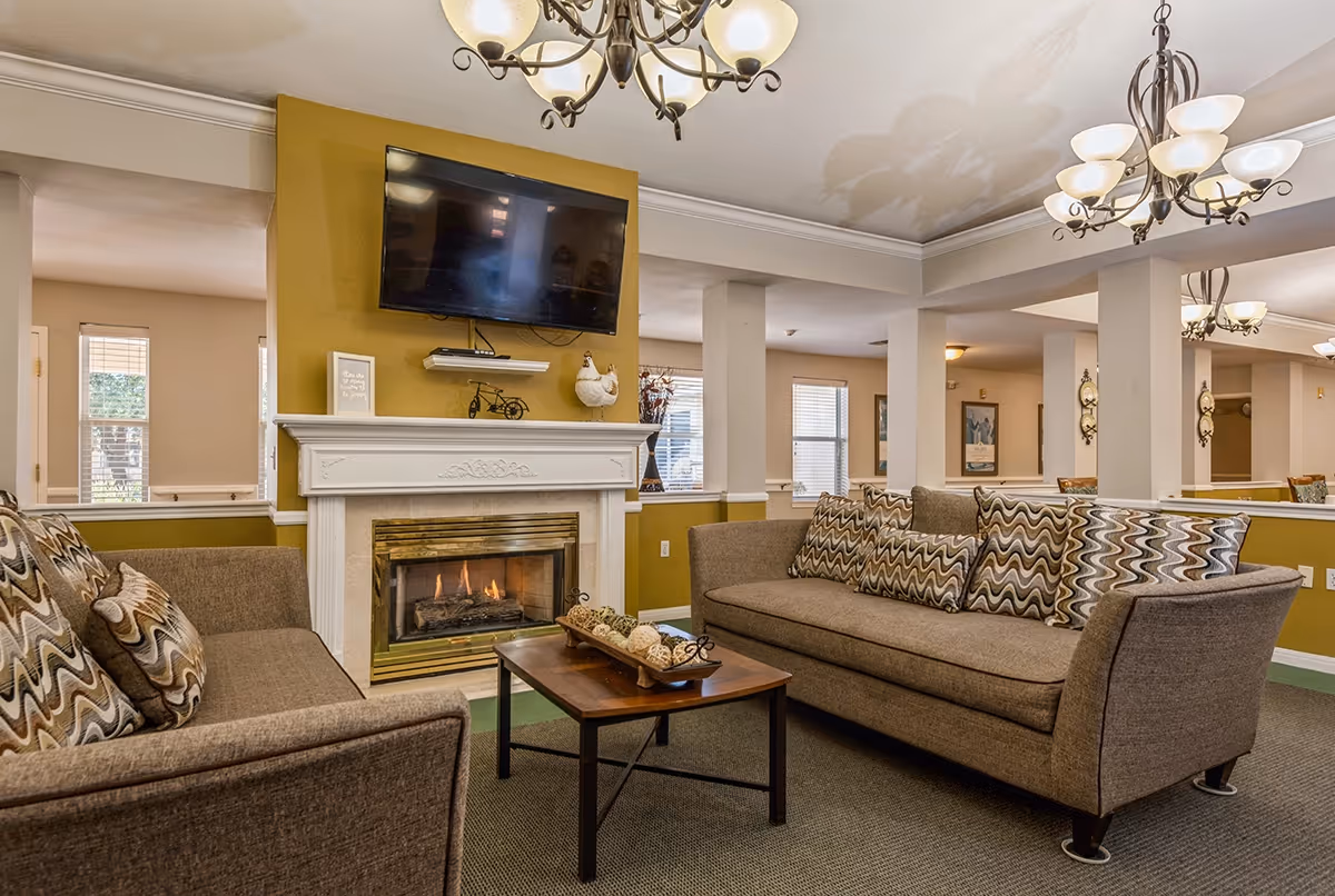 A cozy living room area with two brown sofas adorned with patterned cushions facing a wooden coffee table. A fireplace with a white mantel and a mounted flat-screen TV above it is centered on a mustard yellow accent wall. The room is well-lit with multiple windows and decorative chandeliers hanging from the ceiling.
