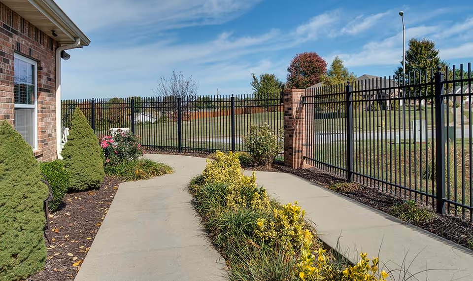 Concrete walkway alongside a brick building with landscaped shrubs and a black metal fence under a blue sky.