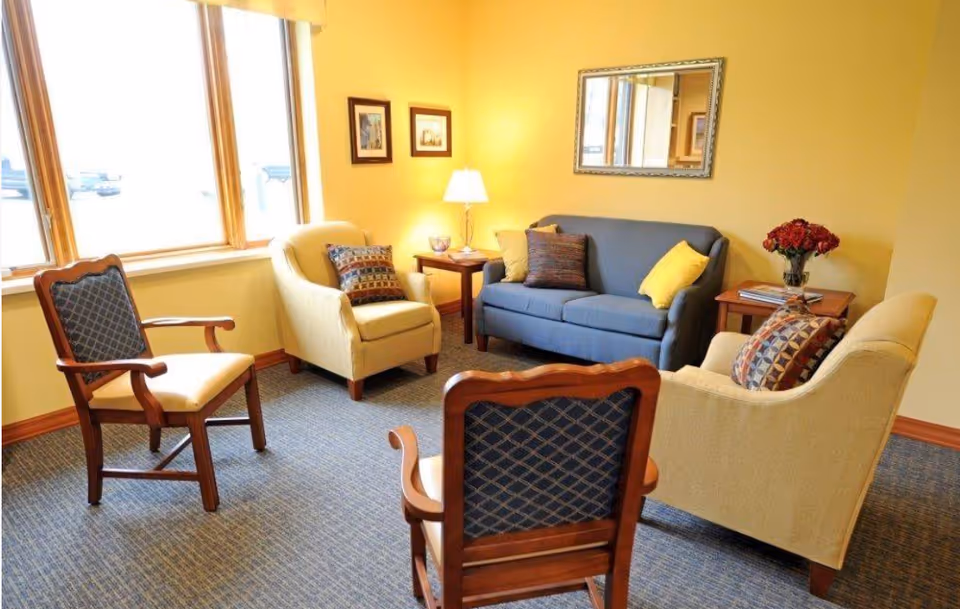 A cozy living room area with a blue sofa, two beige armchairs, and two wooden chairs with blue patterned cushions arranged around a small wooden table with a lamp. The walls are painted yellow and decorated with framed pictures and a mirror. A vase with red flowers sits on a side table.