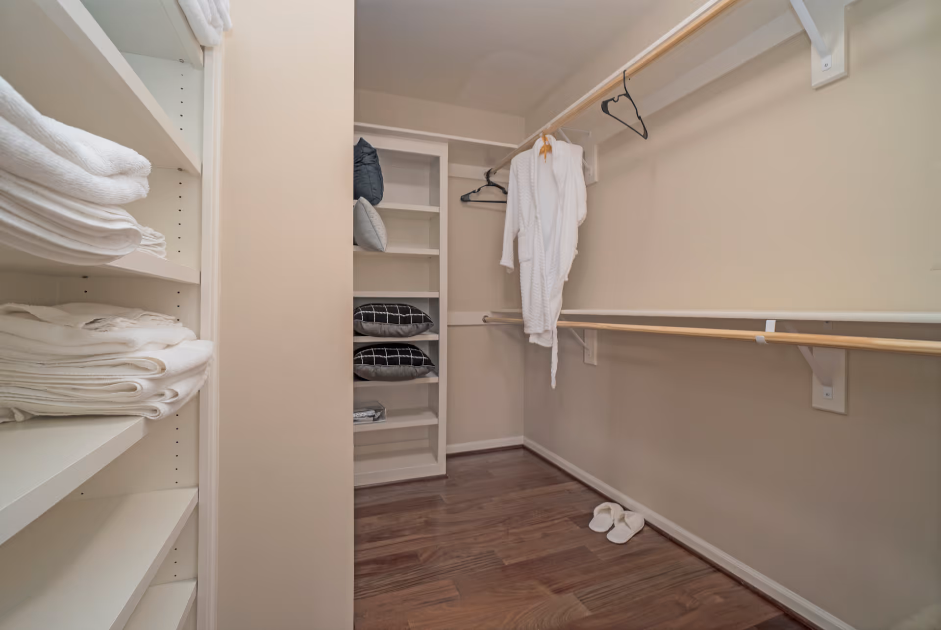 Walk-in closet with built-in shelves holding folded towels and pillows, a hanging white robe, and slippers on a hardwood floor.