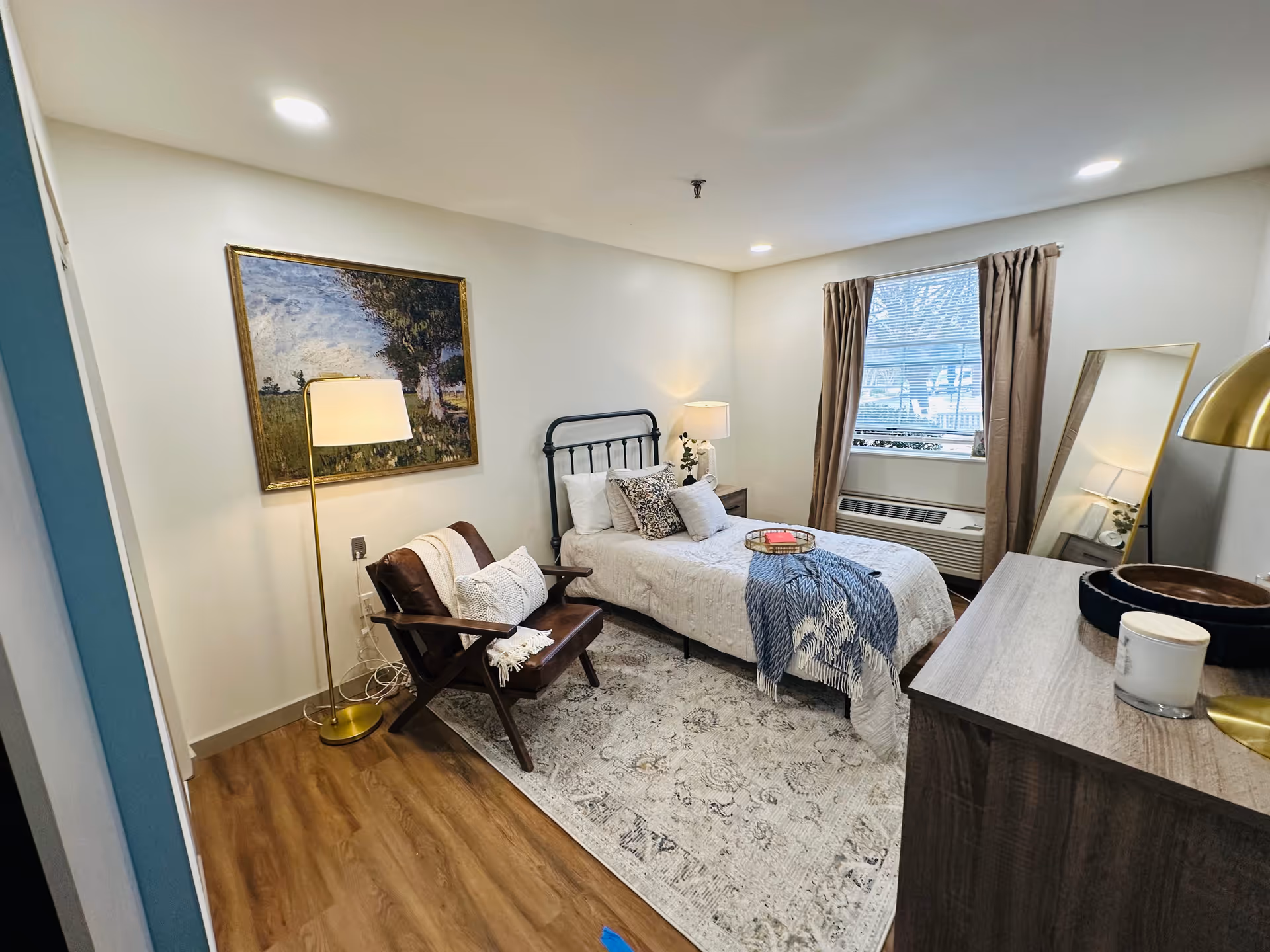 A cozy senior living bedroom featuring a single bed with white bedding and decorative pillows, a wooden armchair with cushions and a throw blanket, a floor lamp, a large framed painting on the wall, a window with brown curtains, a full-length mirror leaning against the wall, and a wooden dresser with decorative items on top. The room has wood flooring and a patterned area rug.