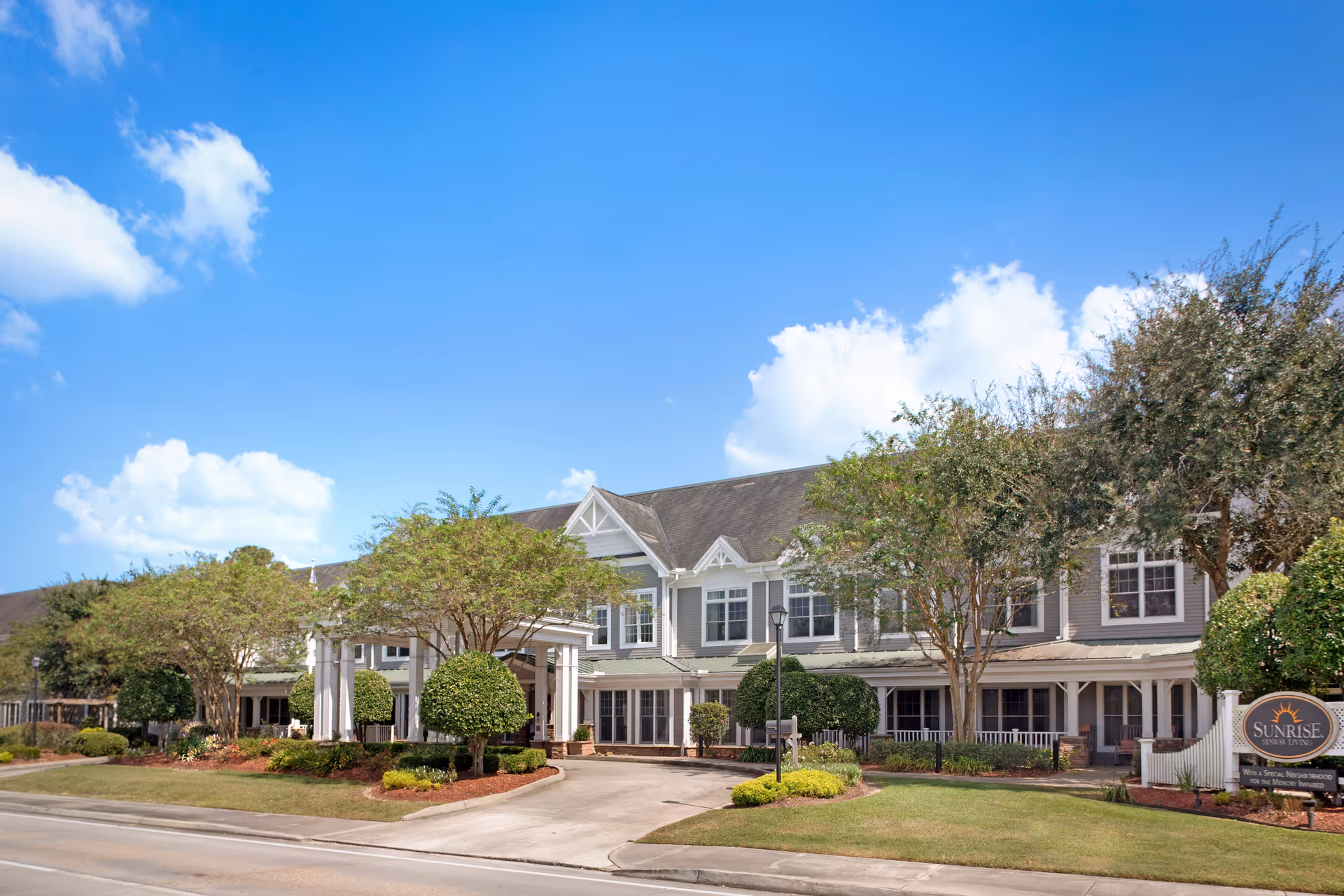 Exterior view of Sunrise at Siegen senior living facility showing a large two-story building with a covered entrance, surrounded by neatly trimmed bushes and trees under a clear blue sky.
