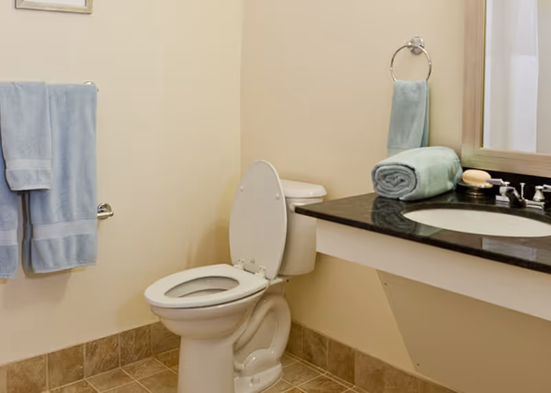 A clean bathroom featuring a white toilet with the lid open, a black countertop with a built-in sink, a rolled-up towel and soap on the counter, a hand towel hanging on a ring, and two blue towels hanging on a wall-mounted towel rack. The floor is tiled and the walls are painted a light beige color.