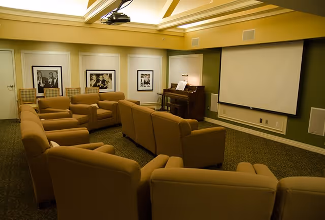 A cozy common room with multiple beige armchairs arranged in rows facing a large white projection screen on a green accent wall. There is a piano with sheet music and a lamp in the corner, and framed black and white photographs on the opposite wall. The ceiling has recessed lighting with a warm glow.