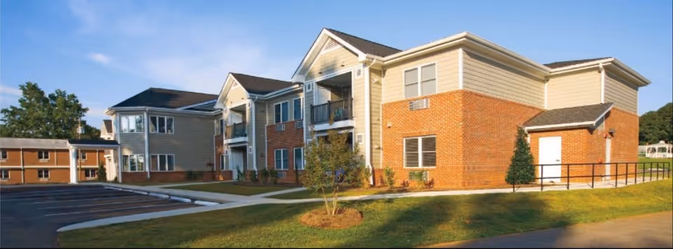 Exterior view of a senior living facility building with brick and beige siding, multiple windows, balconies, a small tree in front, and a parking lot to the left under a clear blue sky.