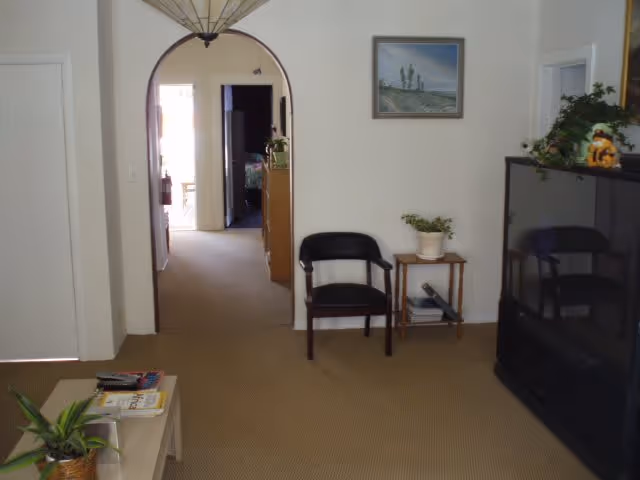 Interior view of a living room area with a beige carpet, a black chair next to a small wooden table with a potted plant, a large TV on the right, and an arched doorway leading to a hallway with more rooms visible in the background.