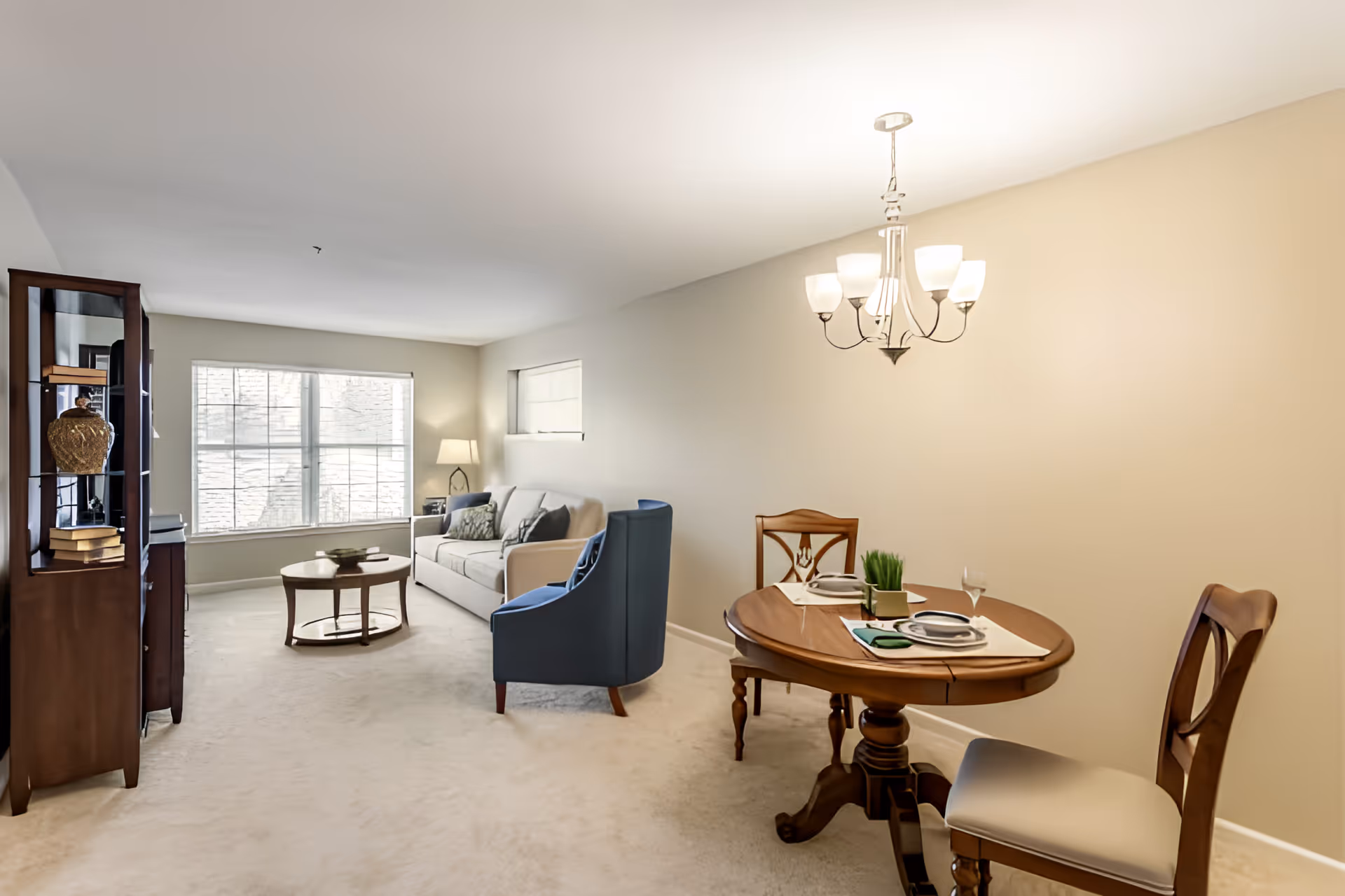 Open living and dining area with a sofa, blue armchair, round wooden coffee and dining tables, and a large window letting in natural light.