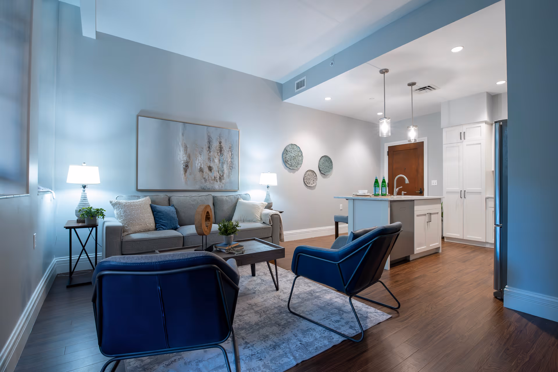 Open-concept living room and kitchen with a sofa, two blue armchairs, coffee table, and a kitchen island with pendant lights.