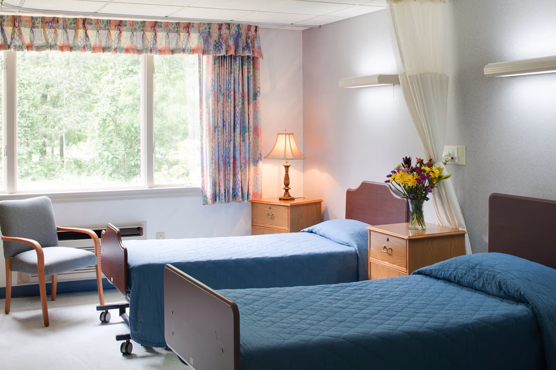 Two hospital-style beds with blue bedspreads in a bright shared resident bedroom with bedside tables, a lamp, flowers, and a large window.