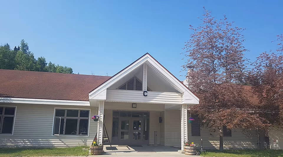 Front exterior view of a single-story building with a peaked roof and a covered entrance marked with the letter C. There are two flower pots with colorful flowers on either side of the entrance, trees with green and reddish leaves, and a clear blue sky above.