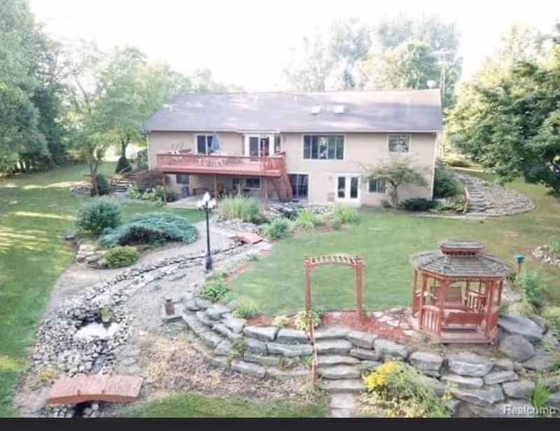 A two-story house with a wooden balcony and stairs leading down to a landscaped garden area. The garden features a stone retaining wall, a small wooden gazebo, a wooden archway, a lamppost, and a dry creek bed lined with rocks. The surrounding area is lush with green grass and trees.