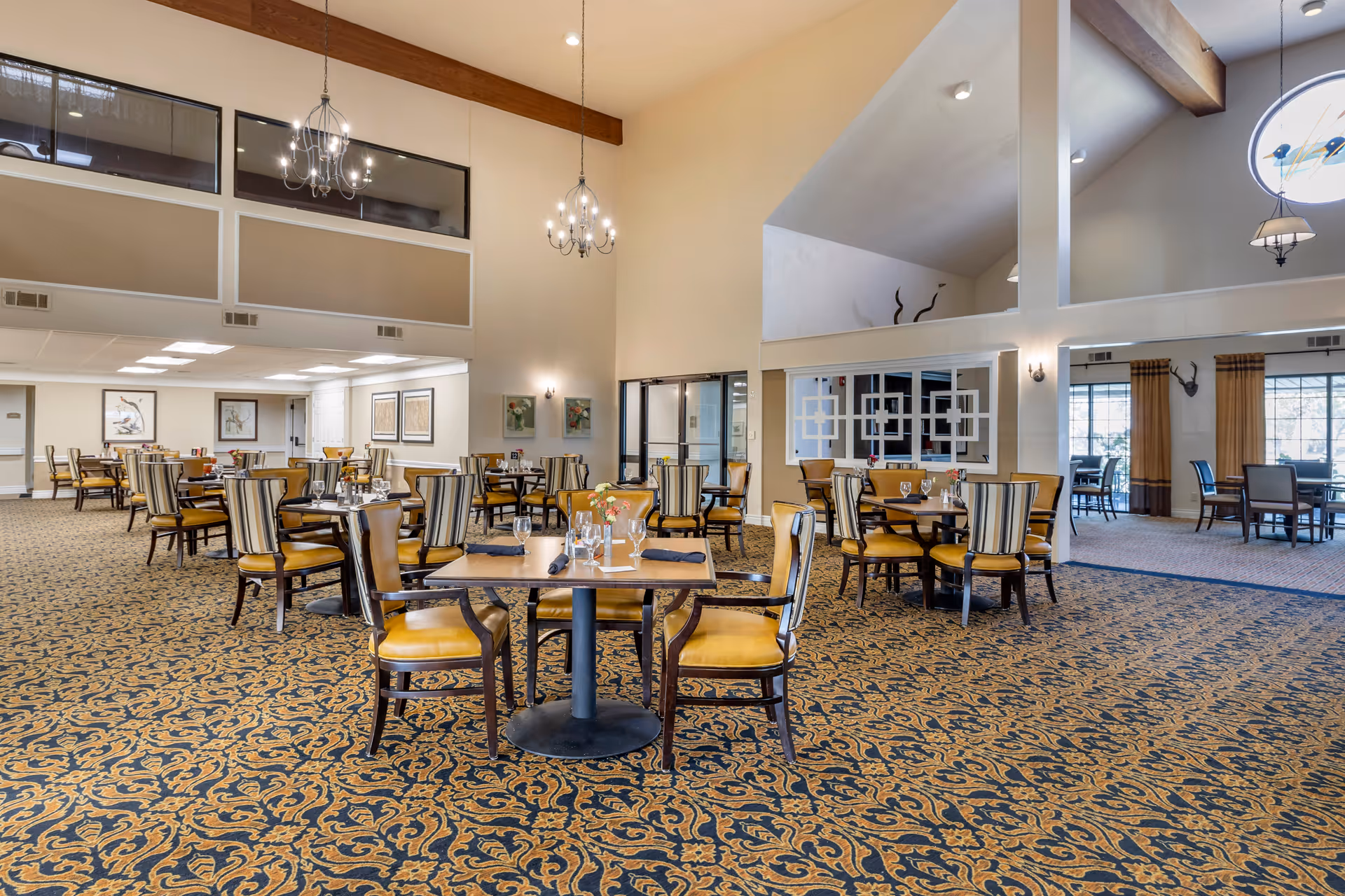 Spacious senior living dining room with multiple round tables and yellow-upholstered chairs beneath chandeliers.
