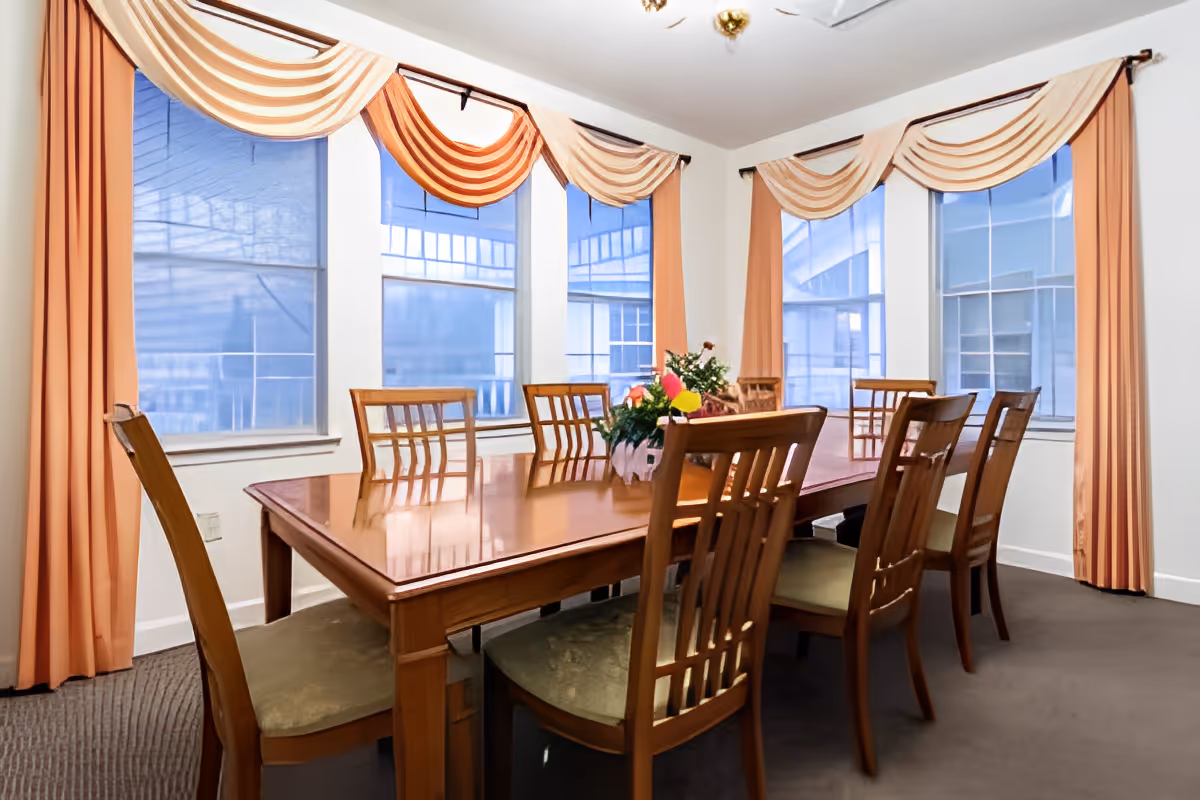 Sunlit dining room with a long wooden table, matching chairs, a floral centerpiece, and large windows with peach drapes.