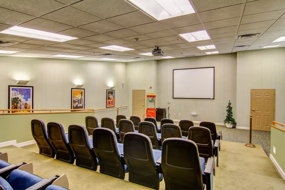 A small theater room with rows of blue cushioned seats facing a blank white projection screen. The room has light green walls with three framed movie posters, a popcorn machine near the screen, and ceiling lights providing illumination.
