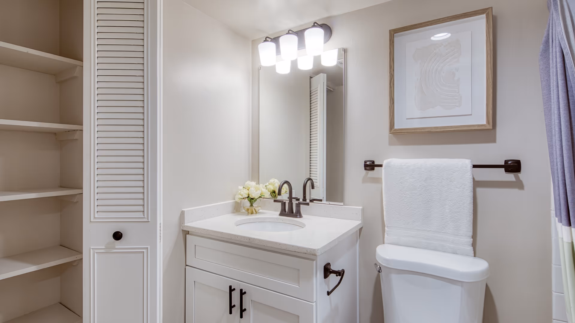 A clean and modern bathroom featuring a white vanity with a sink, a large mirror with three overhead lights, a white toilet with a towel hanging on a rack above it, a framed abstract artwork on the wall, and an open closet with shelves on the left side.