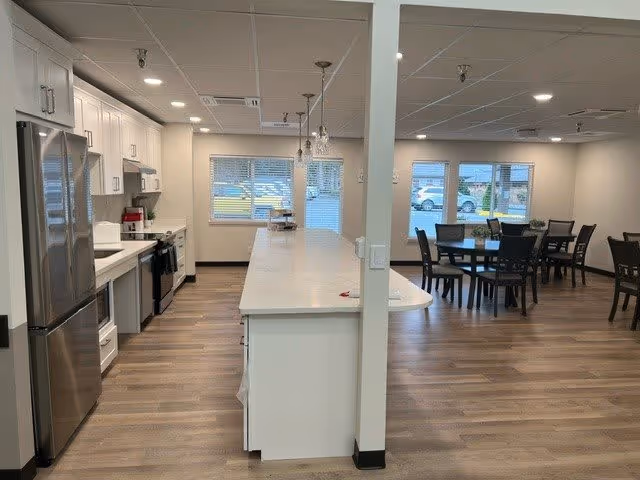 Interior view of a senior community facility showing a modern kitchen area with white cabinets, stainless steel refrigerator, stove, and a long white countertop island. Adjacent to the kitchen is a dining area with multiple dark wood tables and chairs, large windows letting in natural light, and wood flooring throughout.