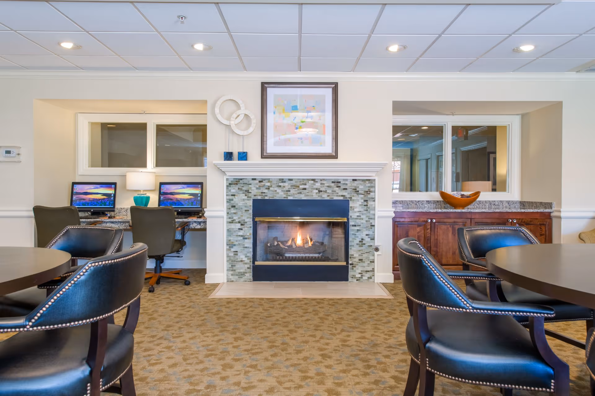 A cozy common area featuring a lit fireplace with a mosaic tile surround, a framed abstract painting above it, and two computer workstations with chairs on the left side. There are round tables with black leather chairs in the foreground, and a wooden cabinet with a decorative bowl on the right side.