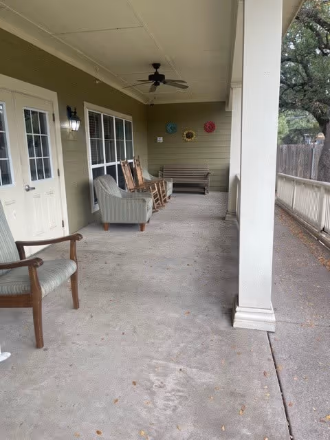 Covered outdoor porch area with several chairs including rocking chairs and a bench. The porch has a ceiling fan, a light fixture, and decorative wall hangings. The area is adjacent to a building with green siding and white trim, and there are trees visible beyond the porch railing.