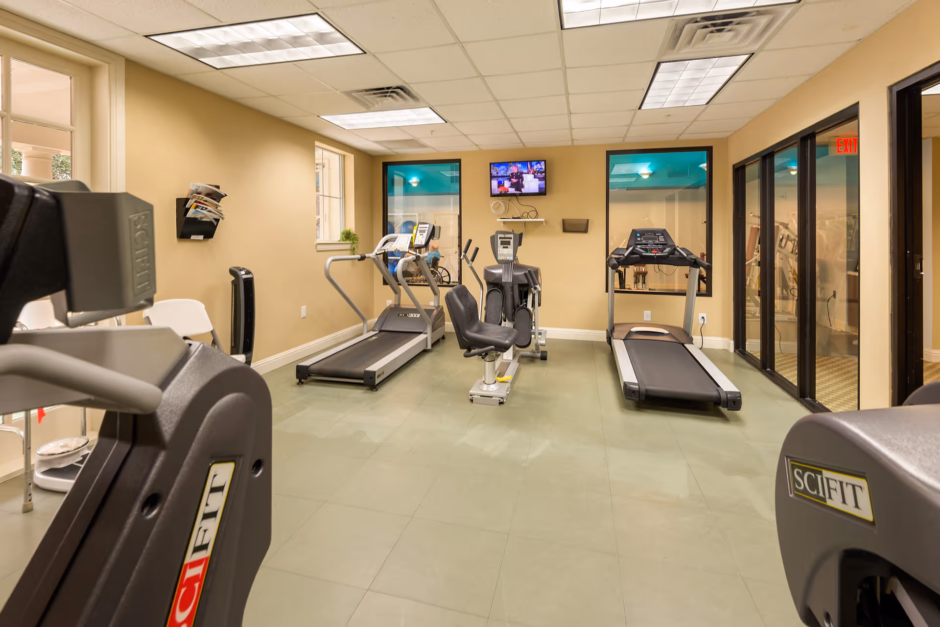 A small fitness room with exercise equipment including two treadmills, a recumbent bike, and an elliptical machine. The room has beige walls, a tiled ceiling with fluorescent lights, and a wall-mounted TV. There are windows and glass doors along one side, and a white chair and magazine rack are visible on the left side.