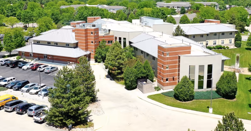 Aerial view of a senior living facility with multiple connected buildings featuring brick and light-colored walls, surrounded by green trees and lawns. There is a parking lot with several cars parked on the left side of the image.