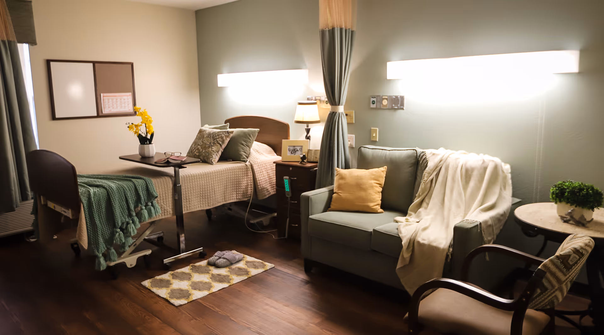 A cozy nursing facility room with a single bed covered in beige and green blankets, a small bedside table with a lamp and framed photo, a green loveseat with a yellow pillow and a white throw blanket, a small round table with a potted plant, and a cushioned chair. The room has wooden flooring and soft lighting from wall-mounted fixtures.