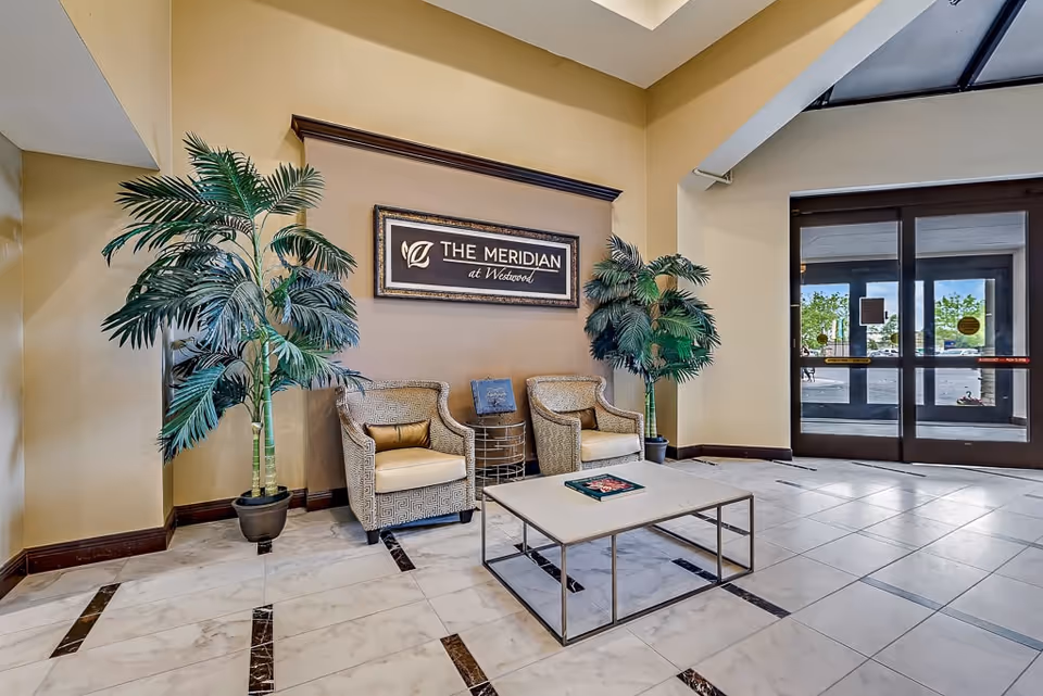 Lobby area of The Meridian at Westwood featuring two patterned armchairs with cushions, a low rectangular coffee table with a book on it, two large potted plants on either side of a framed sign on the wall that reads 'The Meridian at Westwood', and glass double doors leading outside.