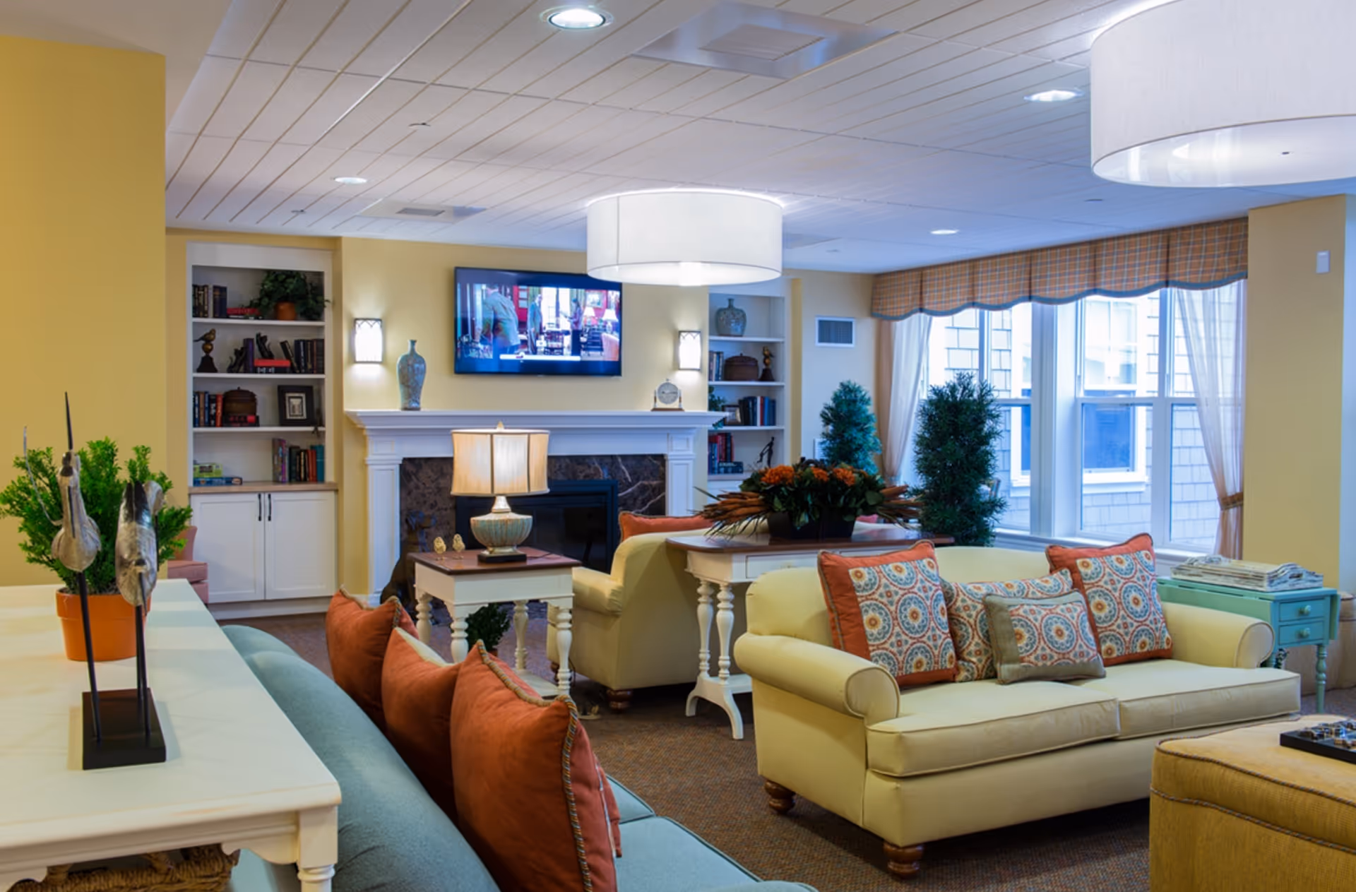 A cozy living room in a senior living facility with yellow walls, a blue sofa with orange pillows, a yellow loveseat with patterned pillows, a fireplace with a mounted TV above it, built-in bookshelves, large windows with curtains, and decorative plants and lamps.