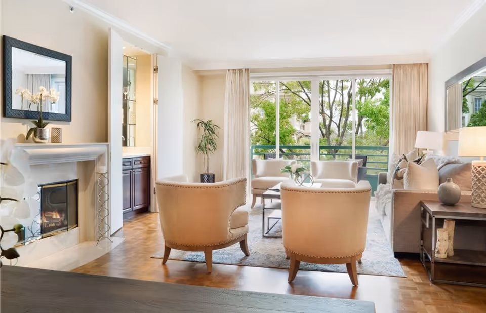 Bright and spacious living room with large windows showing green trees outside. The room features a fireplace with a mirror above it, two beige armchairs, a sofa with cushions, a coffee table with a decorative plant, a side table with a lamp, and light-colored curtains.