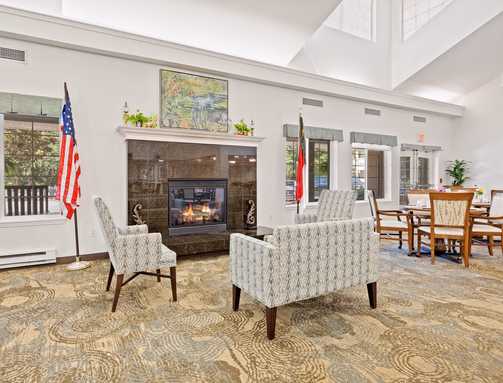 A bright and spacious common area with a fireplace surrounded by dark tiles. In front of the fireplace are two patterned armchairs and a matching loveseat. The room has large windows with green valances, an American flag and another flag on stands, a painting above the fireplace, and a dining table with chairs in the background. The ceiling is high with skylights allowing natural light to fill the space.