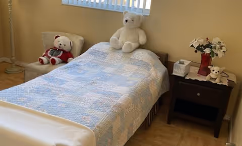 A small bedroom with a single bed covered by a blue and white patchwork quilt. There are two teddy bears on the bed and one teddy bear sitting on a beige chair next to the bed. A dark wooden nightstand beside the bed holds a tissue box, a small teddy bear, and a red vase with white flowers. A window with vertical blinds is above the bed.