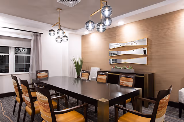 A modern dining room with a long rectangular dark wooden table surrounded by eight chairs with patterned upholstery. The room features a large window with curtains, a decorative wall mirror with geometric shapes, a plant in the corner, and two contemporary pendant light fixtures hanging from the ceiling.