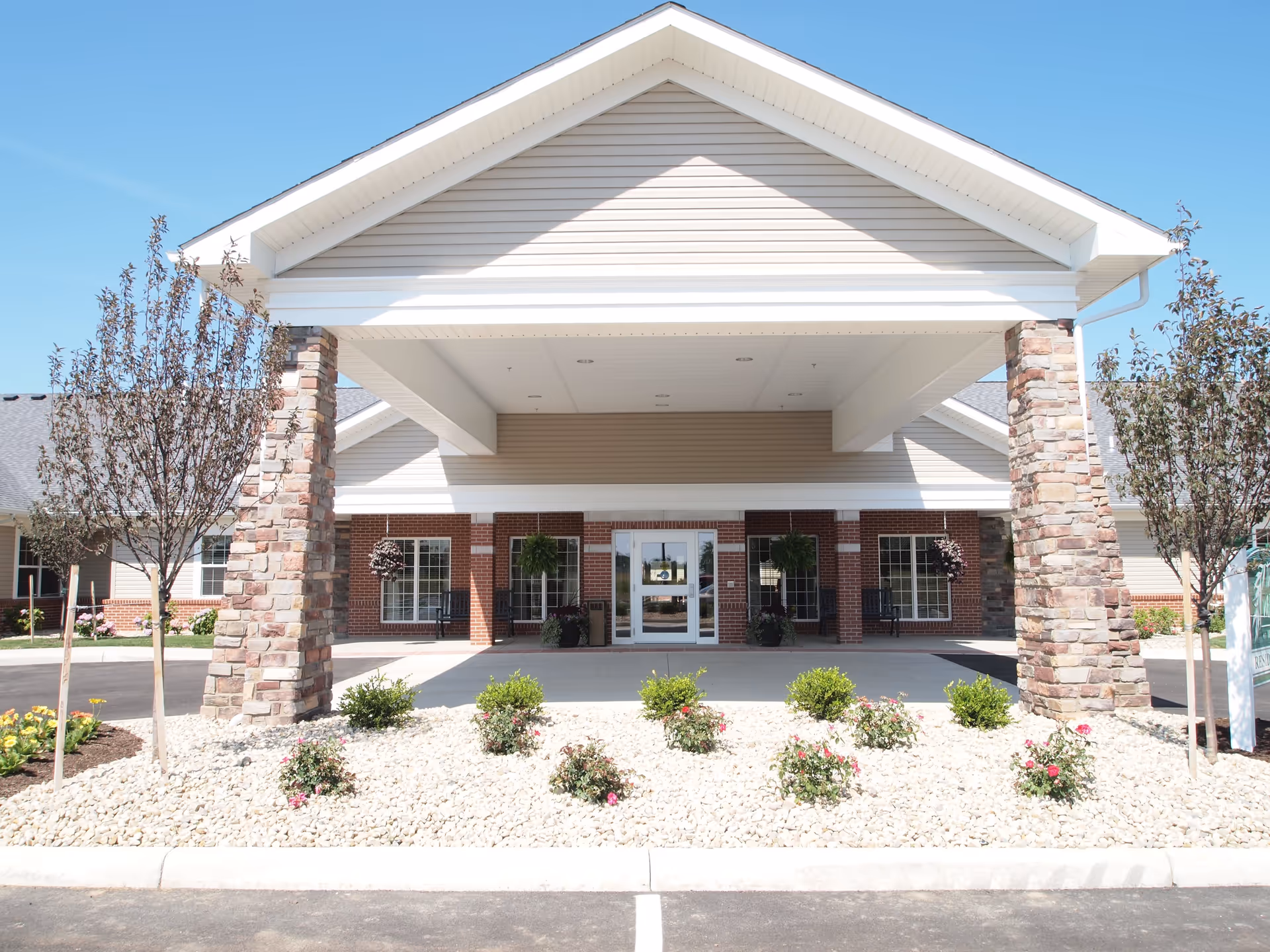 Front exterior view of The Willows at Willard facility with a covered entrance supported by stone pillars, landscaped area with small bushes and flowers, and a clear blue sky.