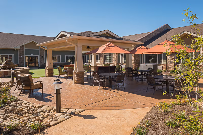 Outdoor patio area at a senior living facility with multiple tables and chairs, some shaded by orange umbrellas, a covered seating area with a ceiling fan, landscaped plants, and a building with stone and siding in the background under a clear blue sky.