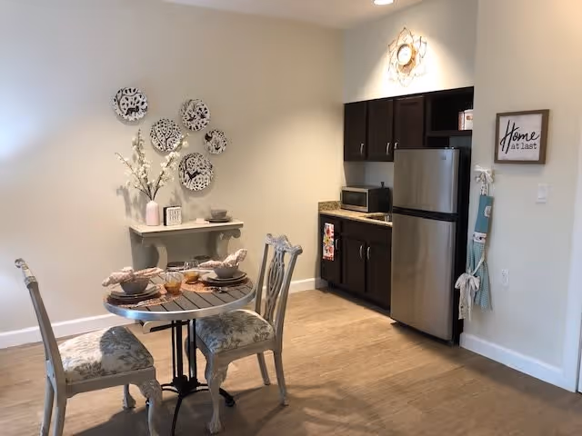 A small dining area with a round table set for two, featuring decorative plates on the wall and a small console table with a vase of flowers. To the right is a kitchenette with dark cabinets, a microwave, and a stainless steel refrigerator. A framed sign on the wall reads 'Home at last.'