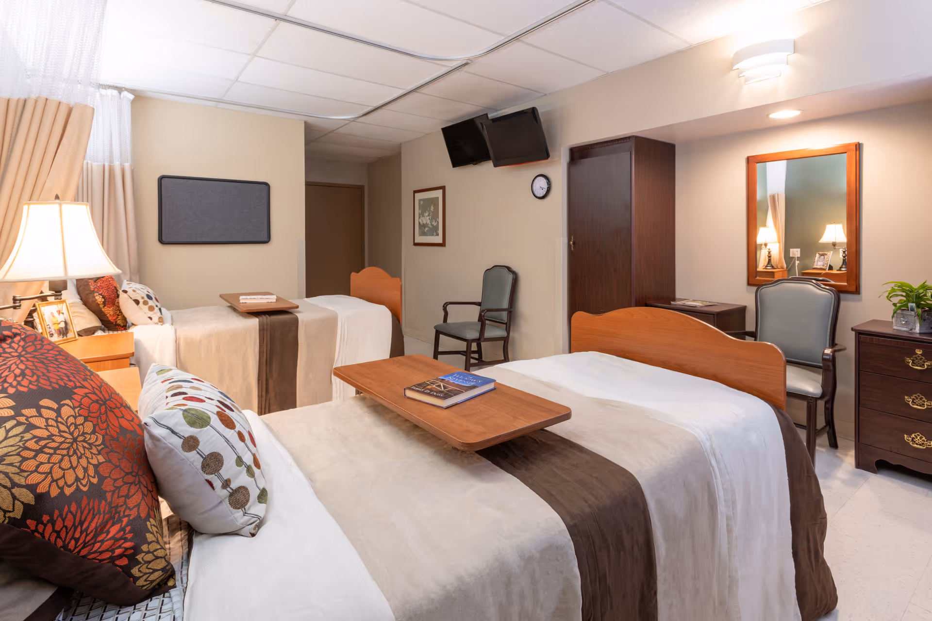 A well-lit bedroom in a senior living facility with two beds, each with a wooden headboard and beige and brown bedding. There are decorative pillows on the beds, a bedside table with a lamp and framed photo, a wooden wardrobe, a wall-mounted TV, a clock, a chair, a dresser with a mirror, and a small plant on the dresser.