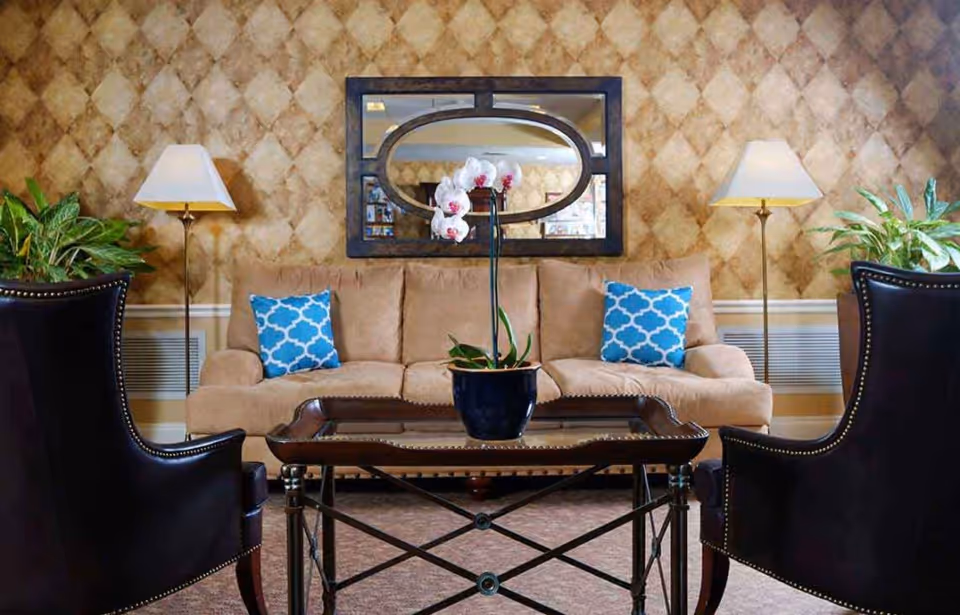 Seating area with a beige sofa, two dark armchairs, a coffee table holding a potted orchid, patterned wallpaper, and floor lamps.
