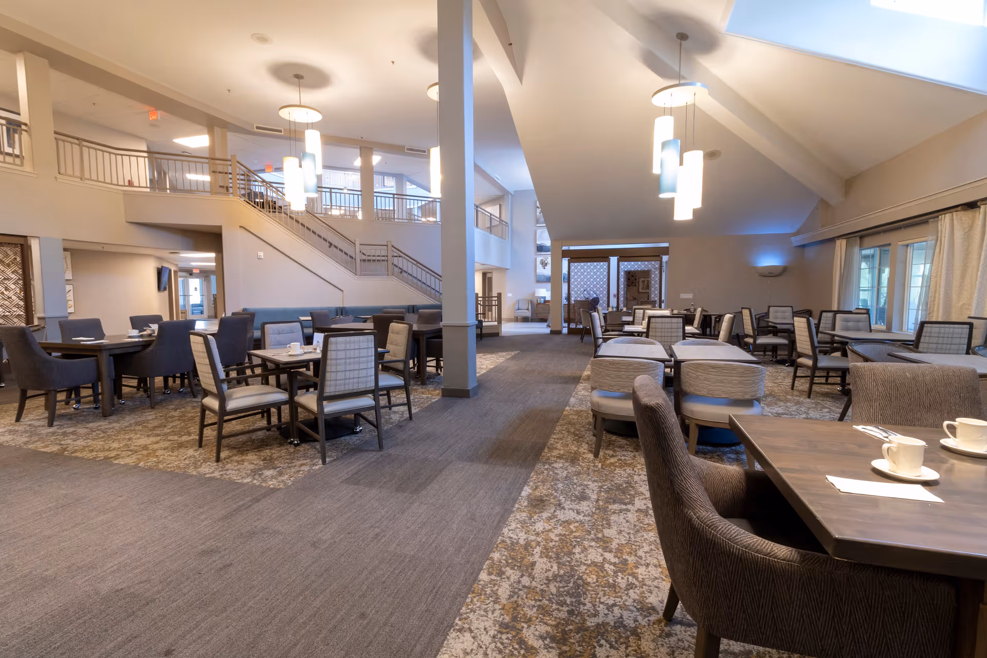 Spacious dining area with multiple tables and chairs arranged neatly, modern pendant lighting hanging from the ceiling, large windows with curtains, and a staircase leading to an upper level in the background.
