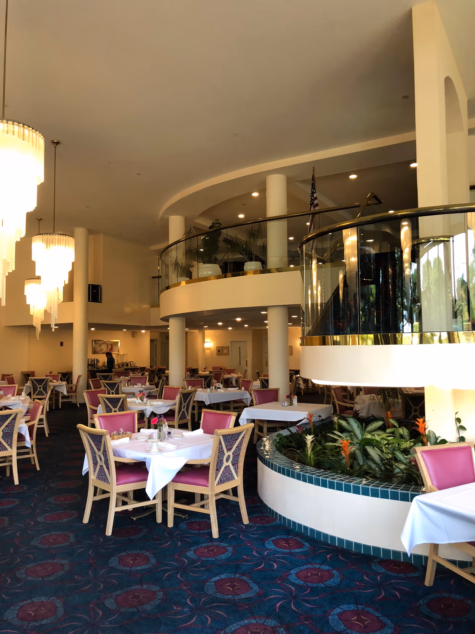 Interior view of a dining room in a senior living facility with multiple tables covered with white tablecloths and set with cups and napkins. The chairs have wooden frames with purple upholstery. There is a curved balcony above with glass railing and several potted plants. Large hanging chandeliers provide lighting, and there is a built-in planter with green plants and orange flowers near the center.