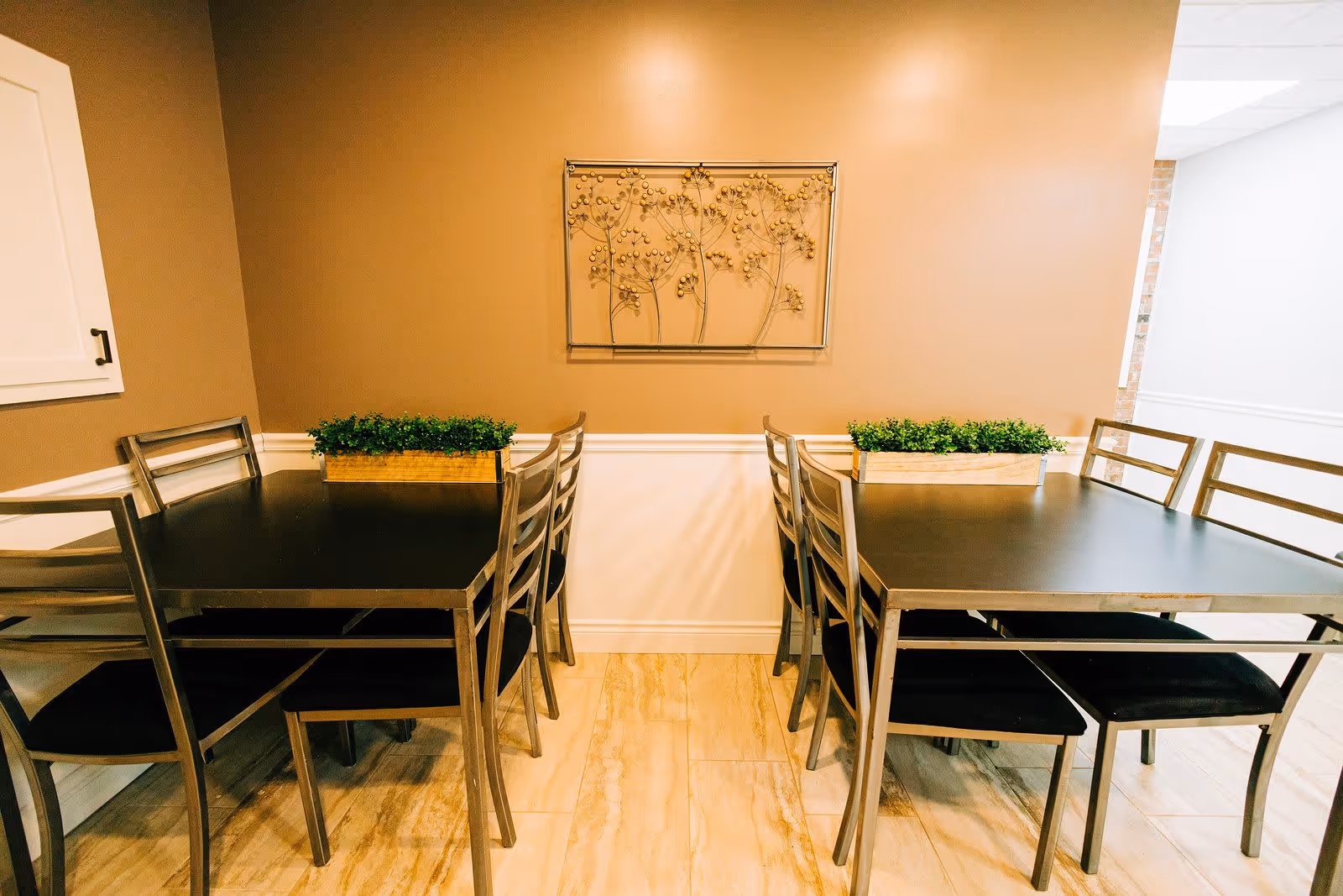 Two black dining tables with metal chairs arranged in a room with beige walls and light wood flooring. Each table has a rectangular planter with green plants. A decorative metal wall art piece featuring a floral design is mounted on the wall above the tables.