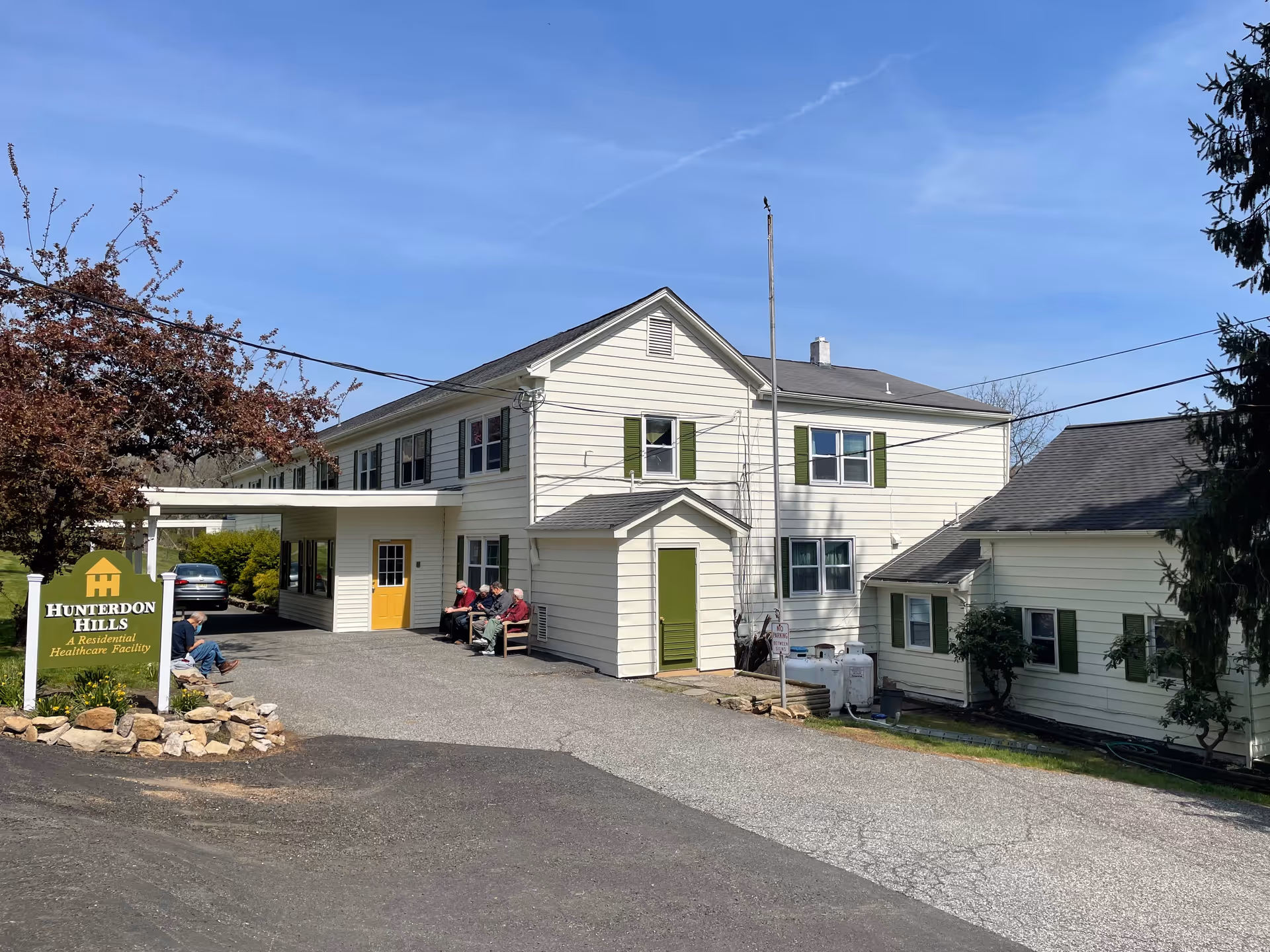Two-story white residential care building with green shutters and a 'Hunterdon Hills' sign at the driveway, with several people seated near the covered entrance.