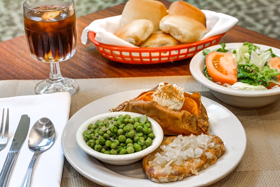 A meal on a wooden table featuring a baked sweet potato topped with sour cream and cinnamon, a serving of green peas in a small bowl, a piece of grilled chicken with chopped onions, a side salad with lettuce and tomato, a basket of dinner rolls, and a glass of iced tea. Silverware is placed on a white napkin beside the plate.