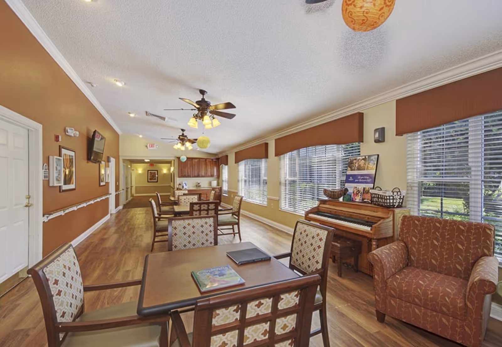 A bright and spacious common area in a senior living facility with several tables and chairs arranged for socializing or activities. The room features large windows with brown valances letting in natural light, ceiling fans with lights, a piano against one wall, and a comfortable armchair. The walls are painted in warm tones, and there is wood flooring throughout.