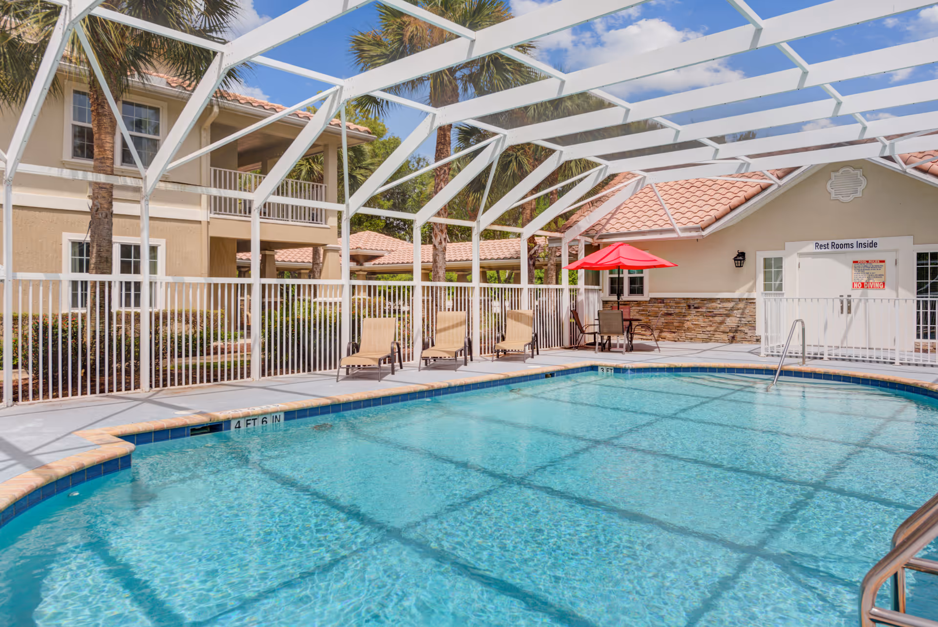 A sunlit outdoor swimming pool with lounge chairs and a red umbrella next to a screened patio and adjacent two-story building.