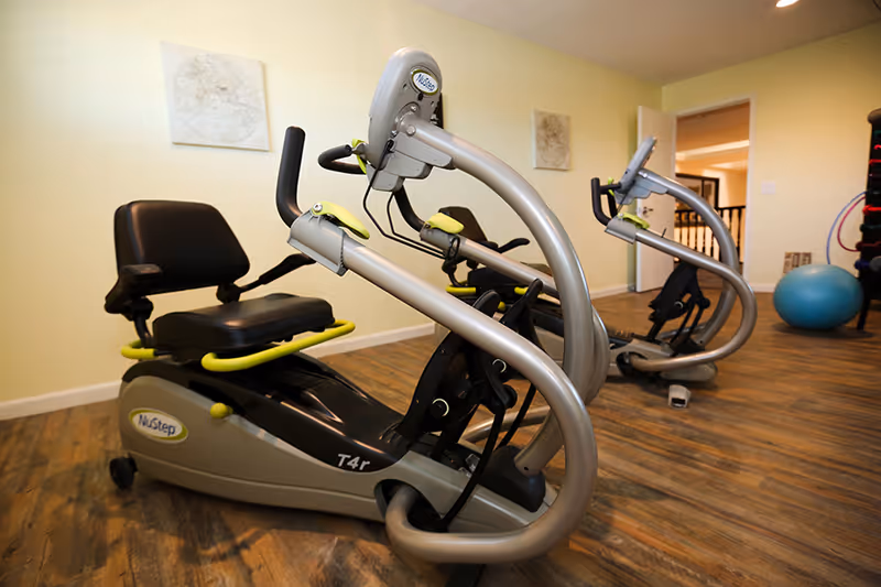 Indoor exercise room with two NuStep T4r recumbent cross trainers on a wooden floor, light yellow walls, and framed artwork. A blue exercise ball and a rack with weights are visible in the background.