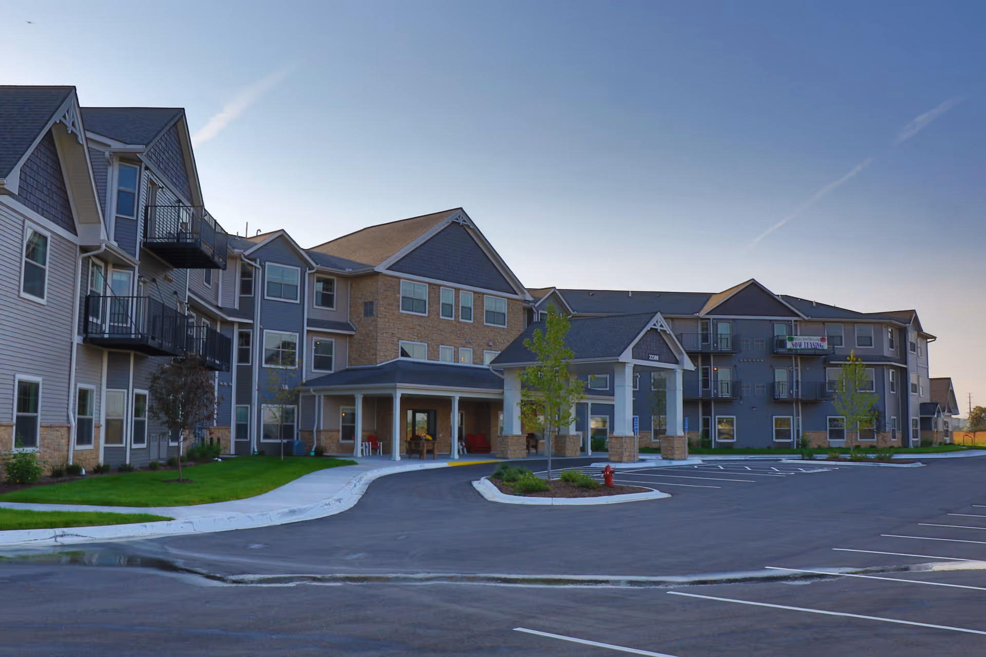 Exterior view of a multi-story senior living facility building with a covered entrance, balconies, and a large parking lot in front under a clear sky.