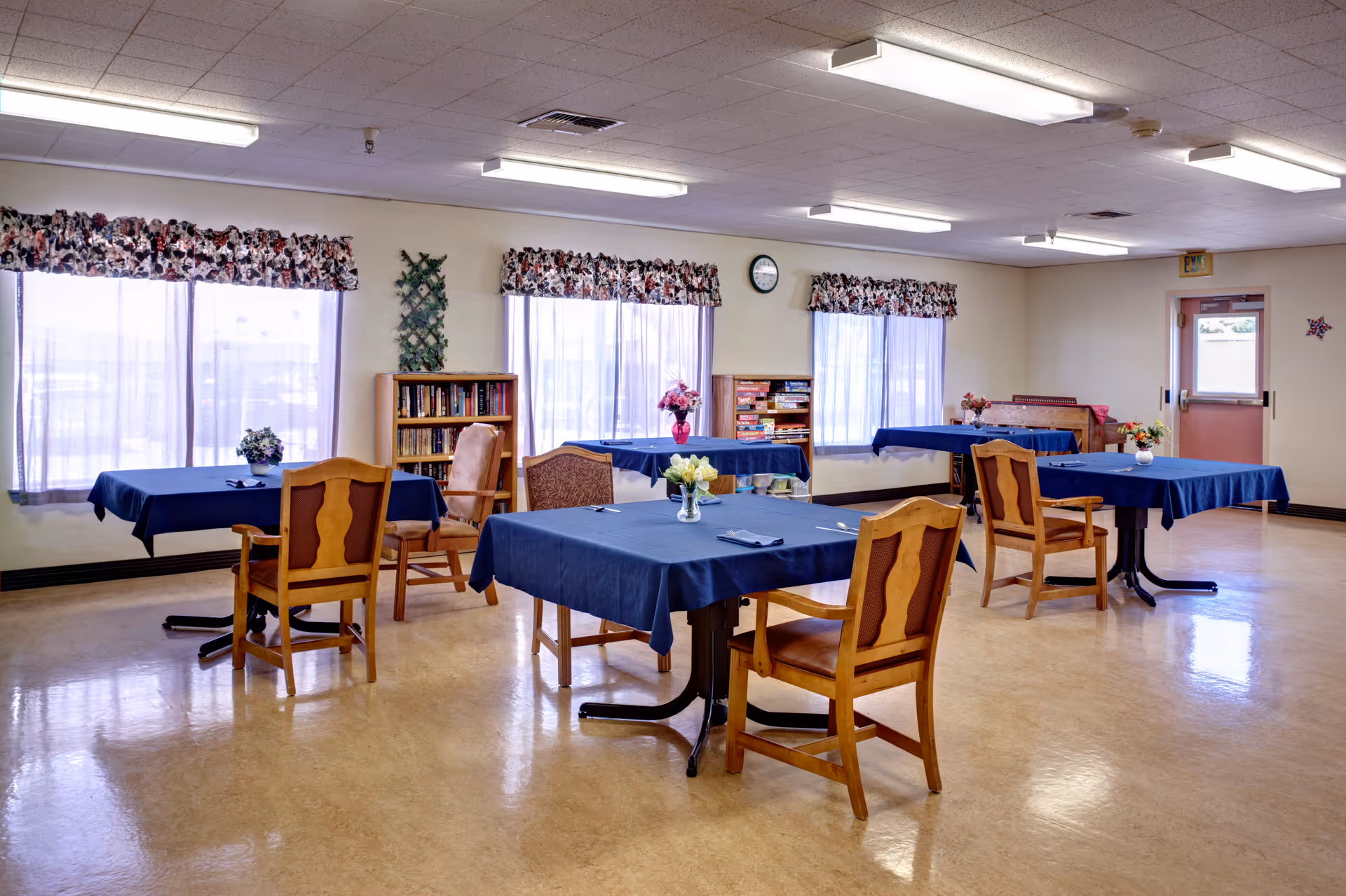 A bright room with several tables covered in blue tablecloths, each surrounded by wooden chairs. The room has large windows with floral valances, bookshelves filled with books and board games, and small flower arrangements on each table. The floor is polished, and fluorescent lights illuminate the ceiling.