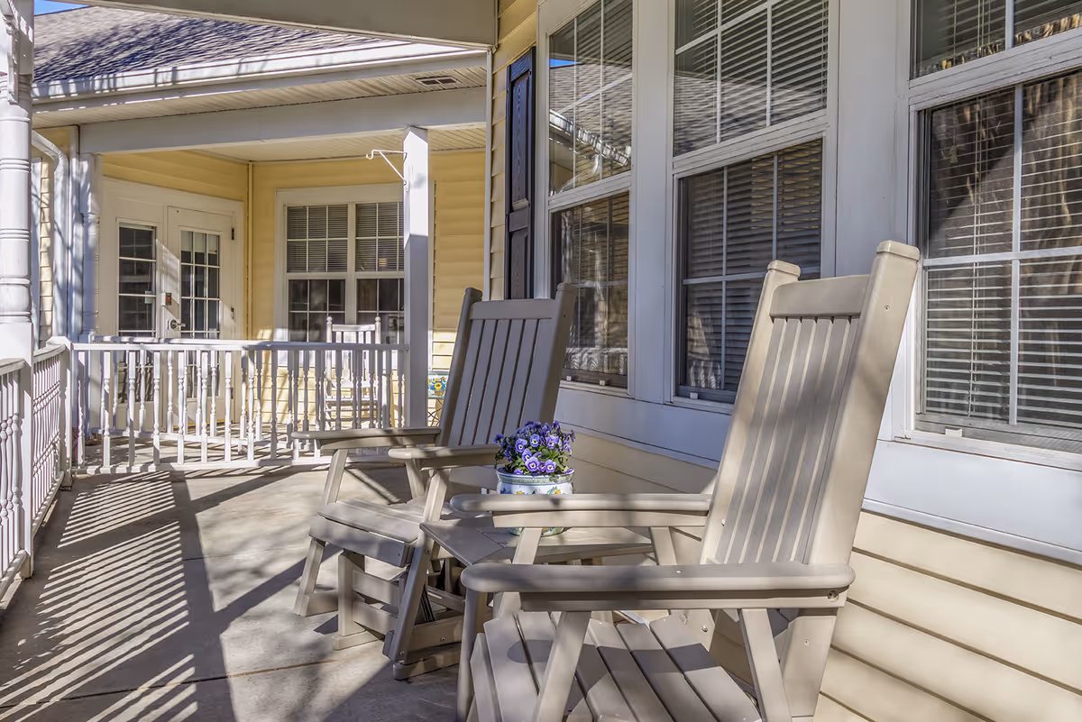 A covered porch area with two beige rocking chairs and a small table holding a pot of purple flowers. The porch has white railings and is attached to a building with yellow siding and multiple windows with white blinds.