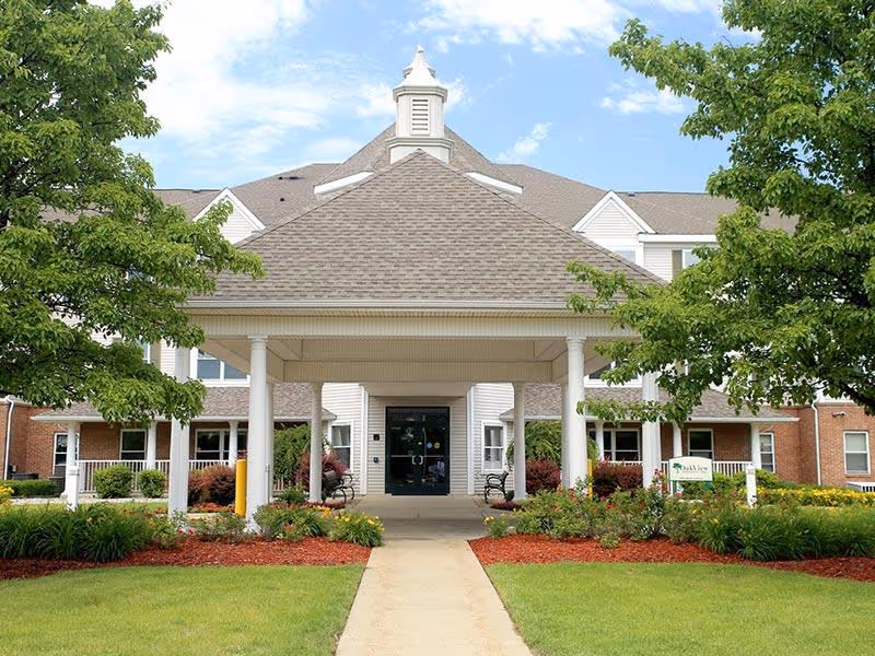 Front exterior view of NorthPointe Woods facility showing a covered entrance with white columns, a pathway leading to the entrance, green lawns, landscaped bushes, and trees under a blue sky with some clouds.